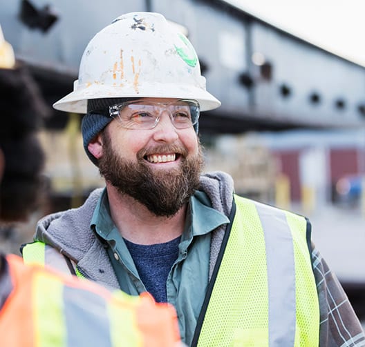 A male construction with a hard hat, safety glasses, and a safety vest worker smiles